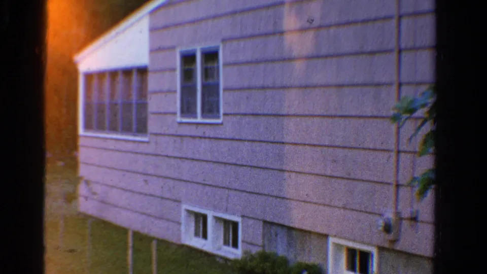 1958 Ontario 8mm frame showing the side of a light-sided house with white windows and basement-level openings above the lawn.