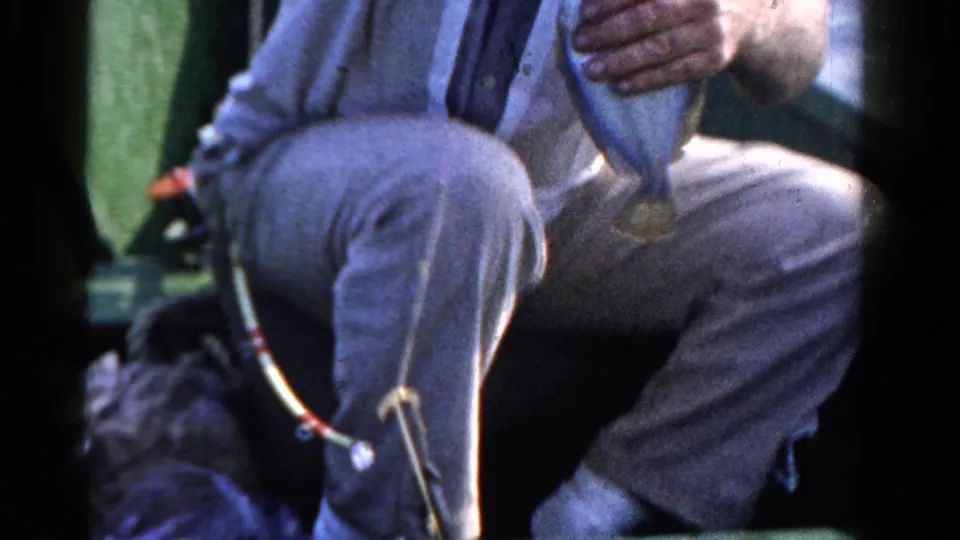A man in casual clothing sits in a small boat and holds up a fish during the Ontario beach trip footage.