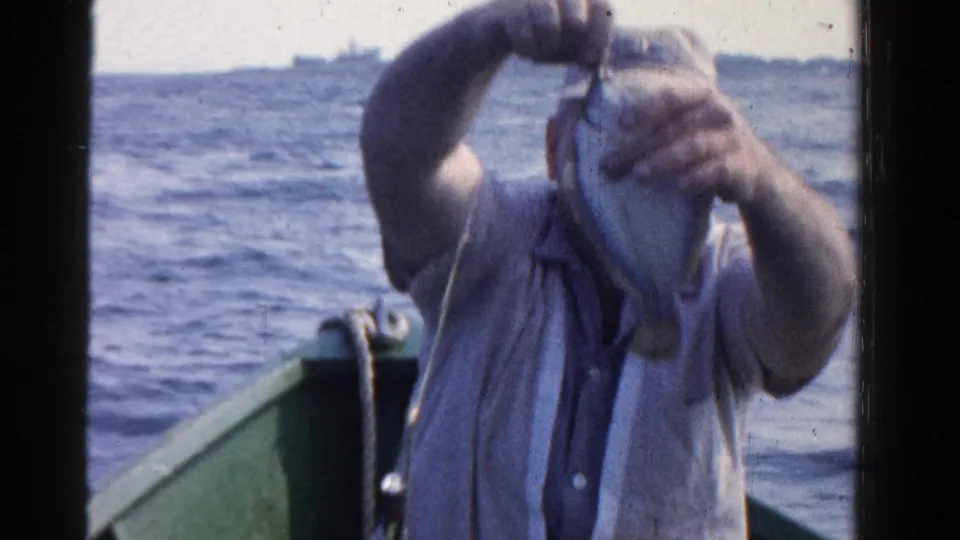 A man aboard a small boat lifts a fish against open water, with distant ships visible in the background.