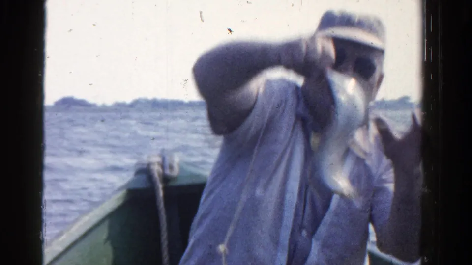 The fisherman displays a silver catch toward the camera in 1958 Ontario home-movie footage from a small boat.