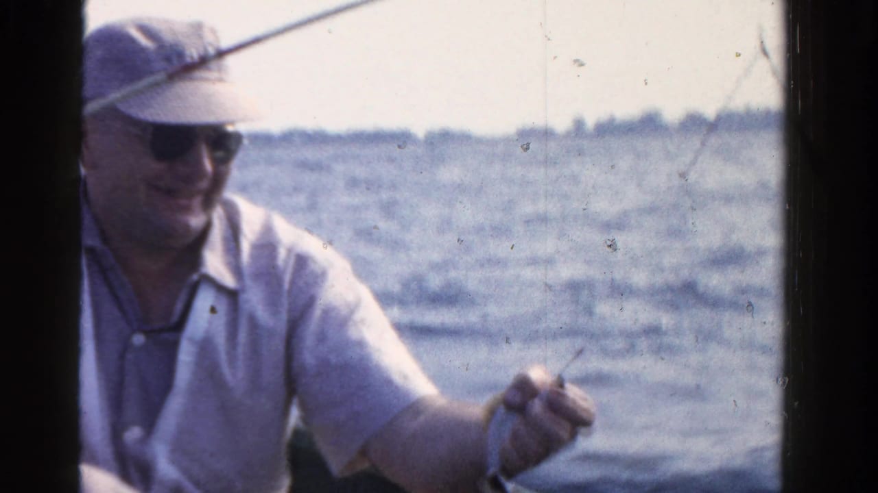 A man wearing sunglasses and a cap smiles while fishing from a small boat on an Ontario lake in 1958.