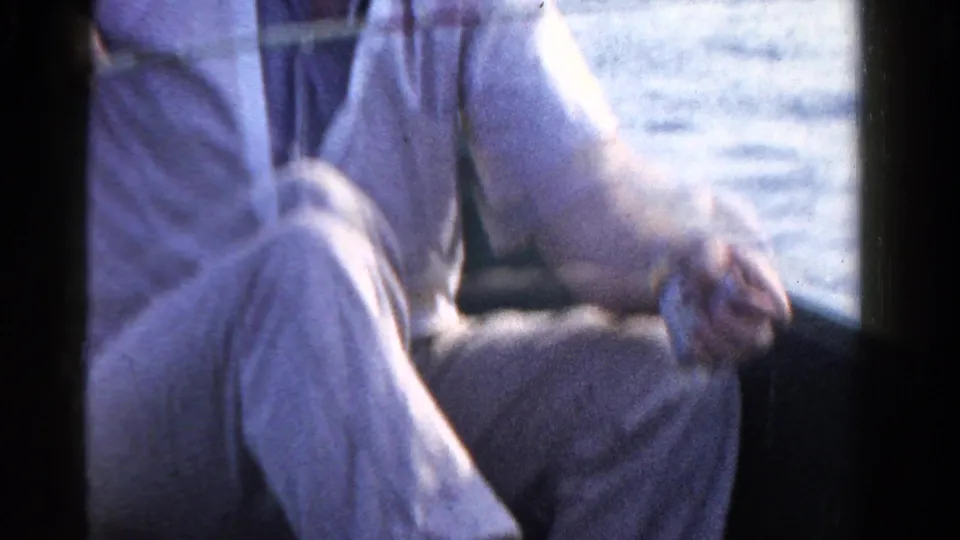 The seated fisherman in light clothing lowers his rod beside the boat, with lake water and shoreline behind him.