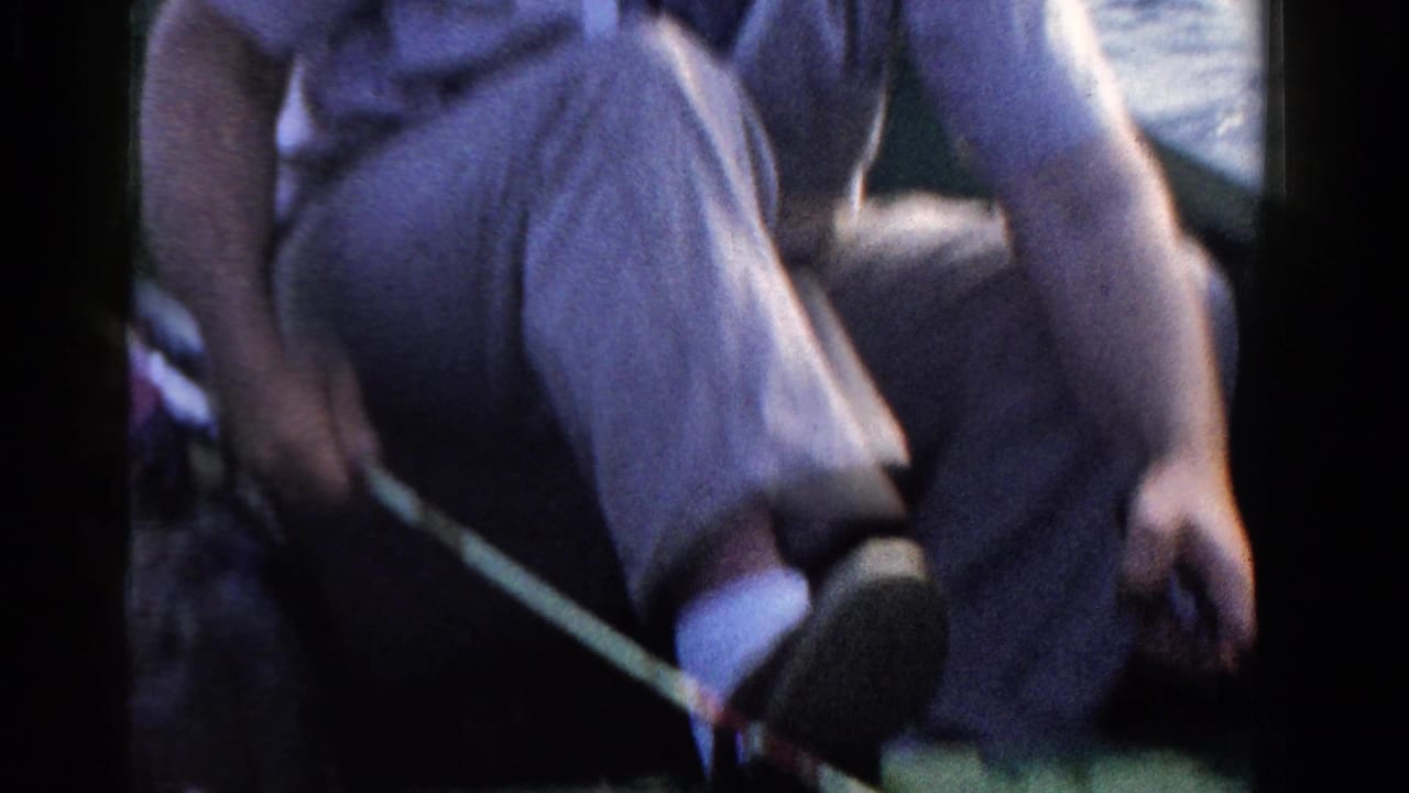 In 1958 Ontario home-movie footage, a seated man in trousers rests in a boat with a fishing rod across the frame and lake water behind him.