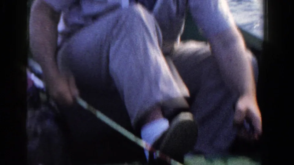 In 1958 Ontario home-movie footage, a seated man in trousers rests in a boat with a fishing rod across the frame and lake water behind him.