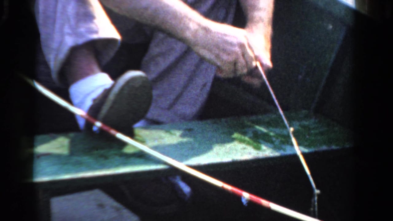 A seated adult man in a boat appears to adjust a fishing line or lure, with his white sock, dark shoe, rod, and green bench visible.