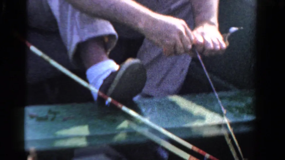 A seated man in 1958 Ontario footage grips a fishing rod and line, framed tightly over the green boat interior with his shoe and sock visible.