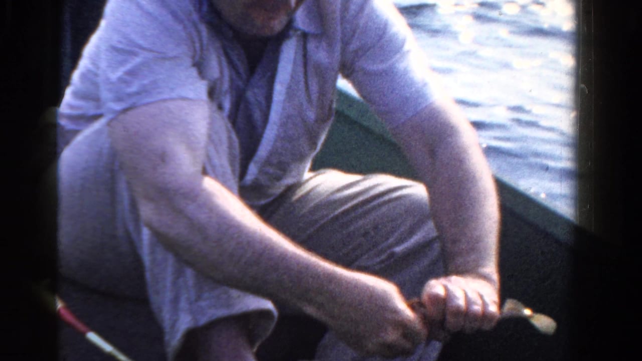 Adult man in 1958 Ontario home-movie footage sits in a small boat and turns a fishing reel beside sunlit water.