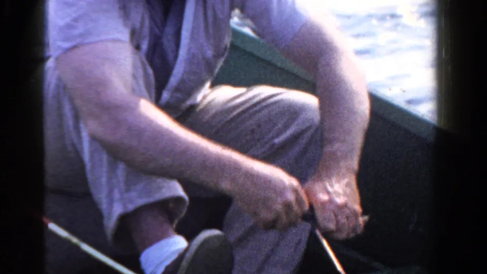 The seated angler remains low in the small boat, with his rod angled across the frame and sunlit lake water beyond.