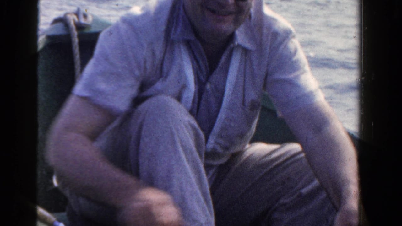 A man in a light shirt and trousers sits crouched in a small boat during 1958 Ontario lake fishing footage.