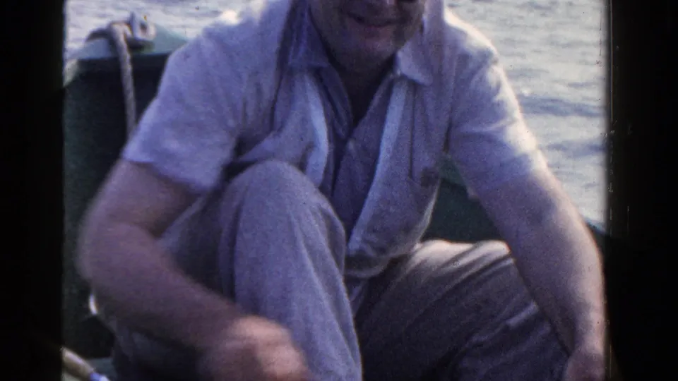 A man in a light shirt and trousers sits crouched in a small boat during 1958 Ontario lake fishing footage.