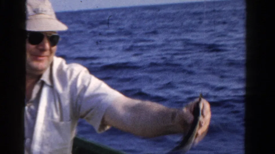 A man in sunglasses and a light hat holds up a small fish from a boat on an Ontario lake in 1958 home-movie footage.