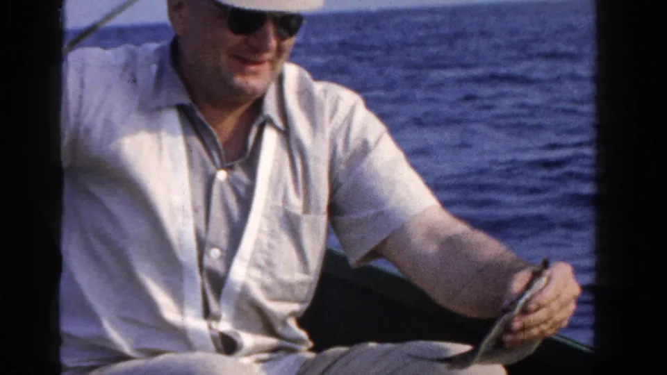 A smiling man in sunglasses and a light shirt holds a small fish from a boat on a lake, matching 1958 Ontario home-movie metadata.