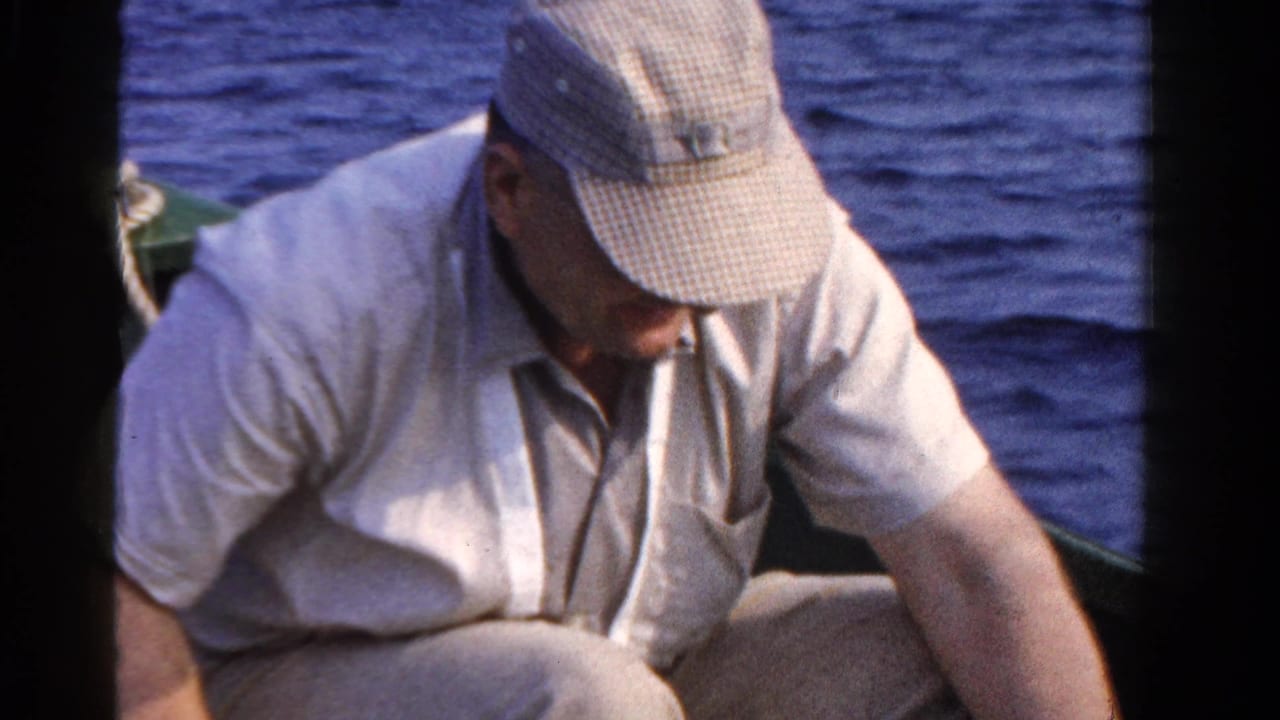 A man wearing a cap and light shirt sits low in a small boat on open water in the 1958 Ontario fishing scene.