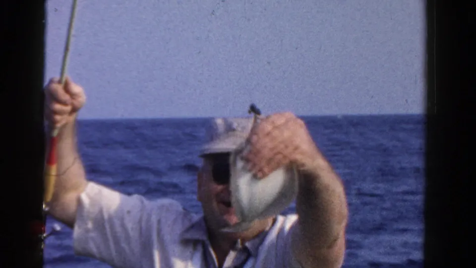 In the Ontario fishing home movie, the man lifts his rod while holding a small fish near his face, with the horizon behind him.