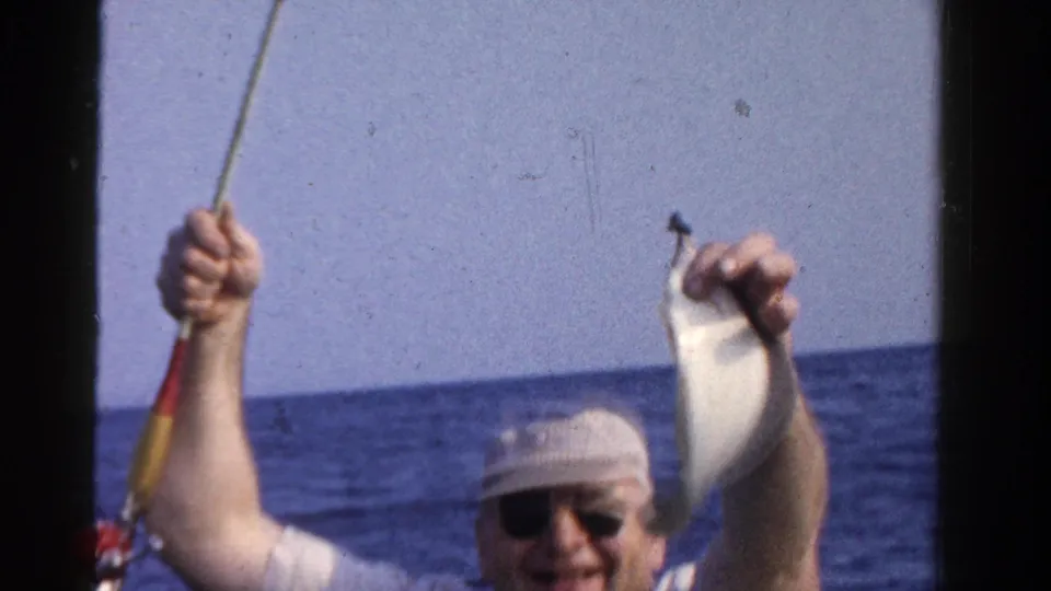 The fisherman smiles toward the camera while holding up a small fish and rod during a 1958 Ontario boat outing.