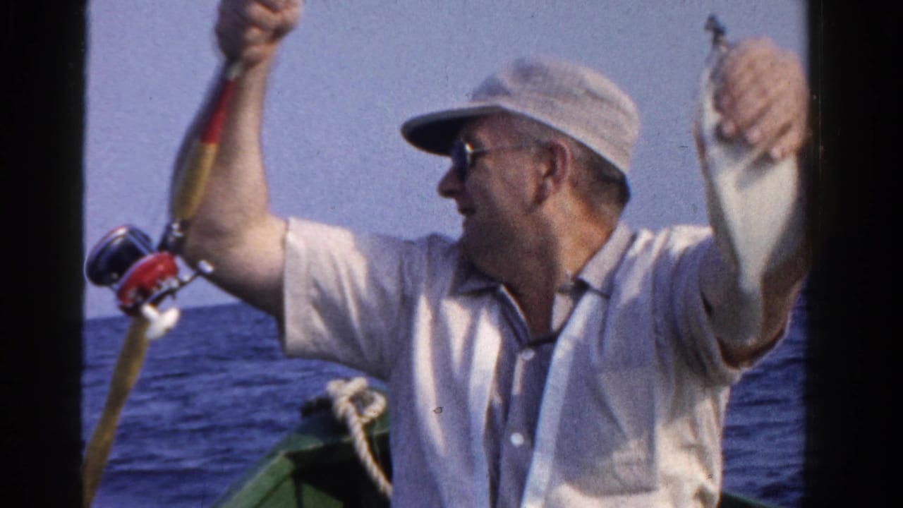 In 1958 Ontario home-movie footage, an older man in a light shirt and cap raises a fishing rod from a small boat on blue water.