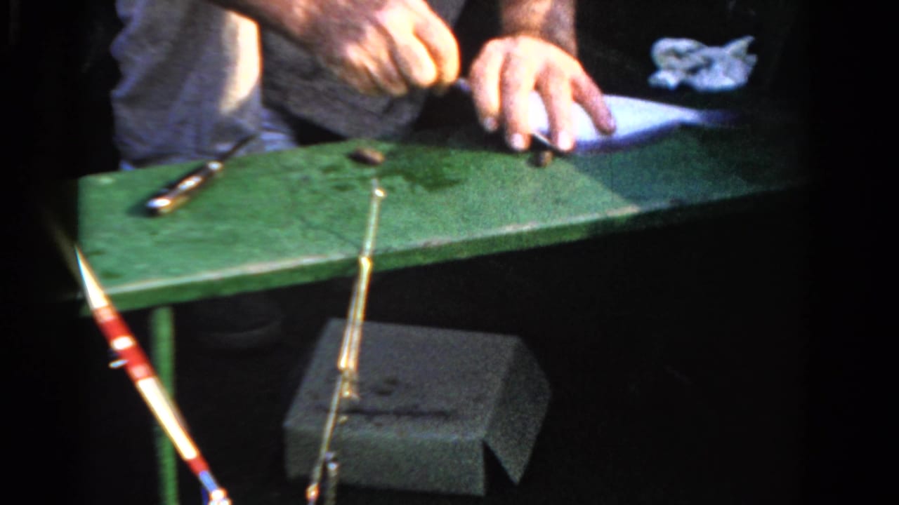 Hands work near a small fish on a green board inside a boat, with a fishing rod leaning across the frame.