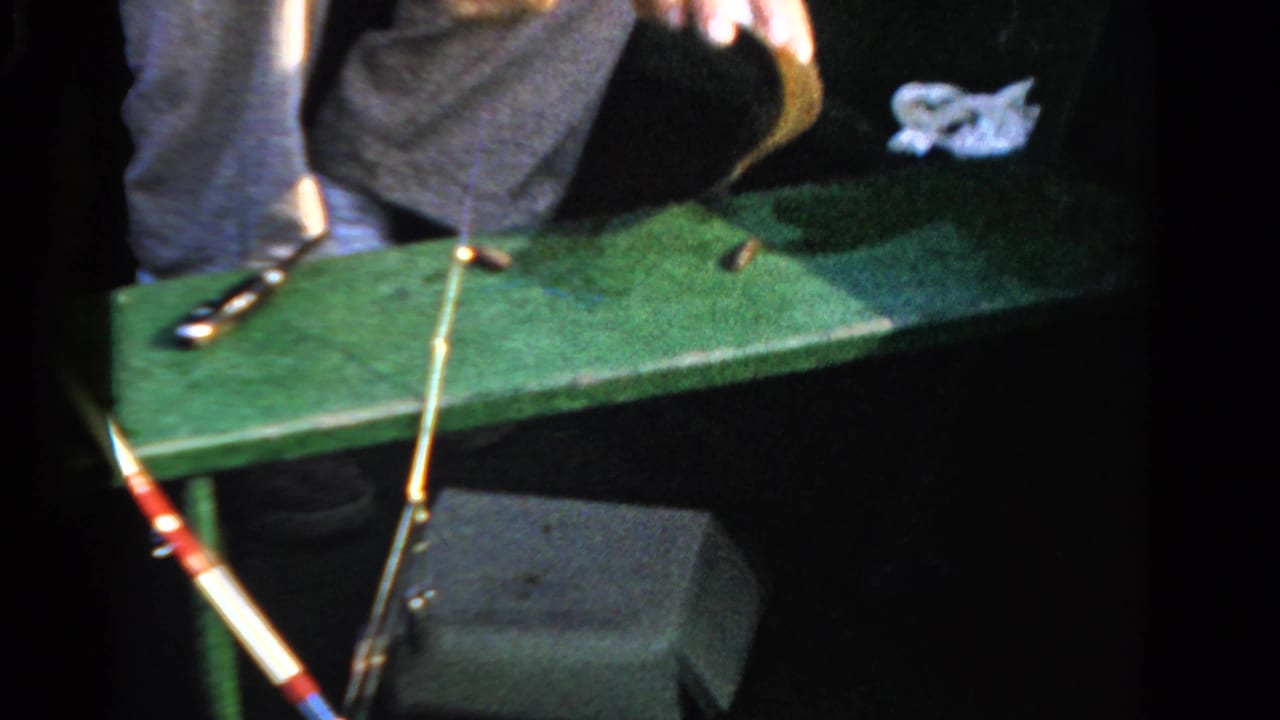 1958 Ontario home-movie frame of a man in a boat leaning over a green board while working near a small fish and rod.