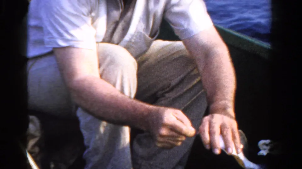 1958 Ontario 8mm still with a seated man in light clothing handling fishing gear or a small fish inside a boat.