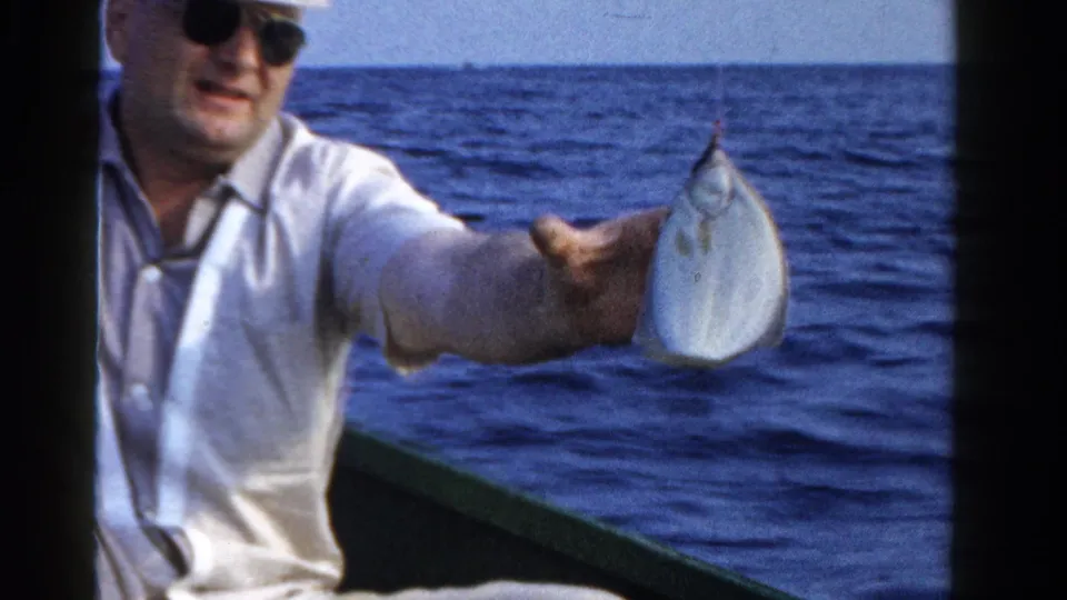 A man in sunglasses holds a small caught fish toward the camera from a boat on open water in 1958 Ontario footage.