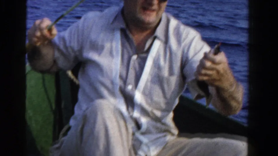 A seated man in a light shirt holds fishing line and gear in a green boat, with blue water visible behind him.