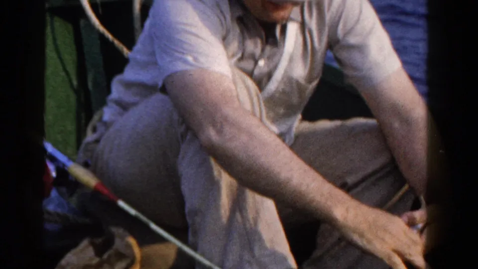 The fisherman bends down inside the small boat, with a rod and rail visible against the water.