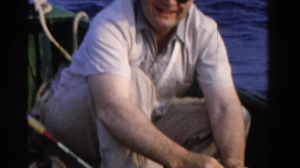 A seated man smiles toward the camera in 1958 Ontario fishing footage, with boat details and water behind him.