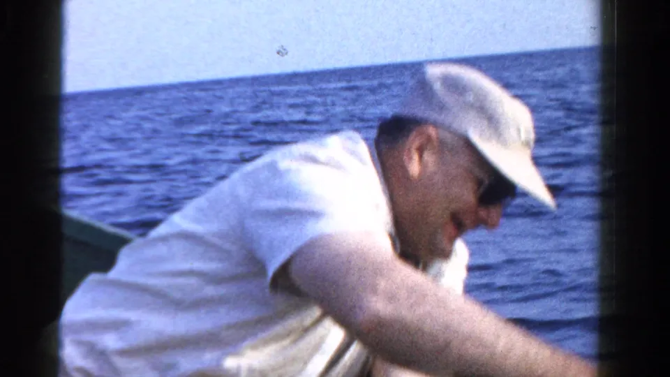 A man in a white cap bends over the side of a small boat on blue water in 1958 Ontario home-movie footage.