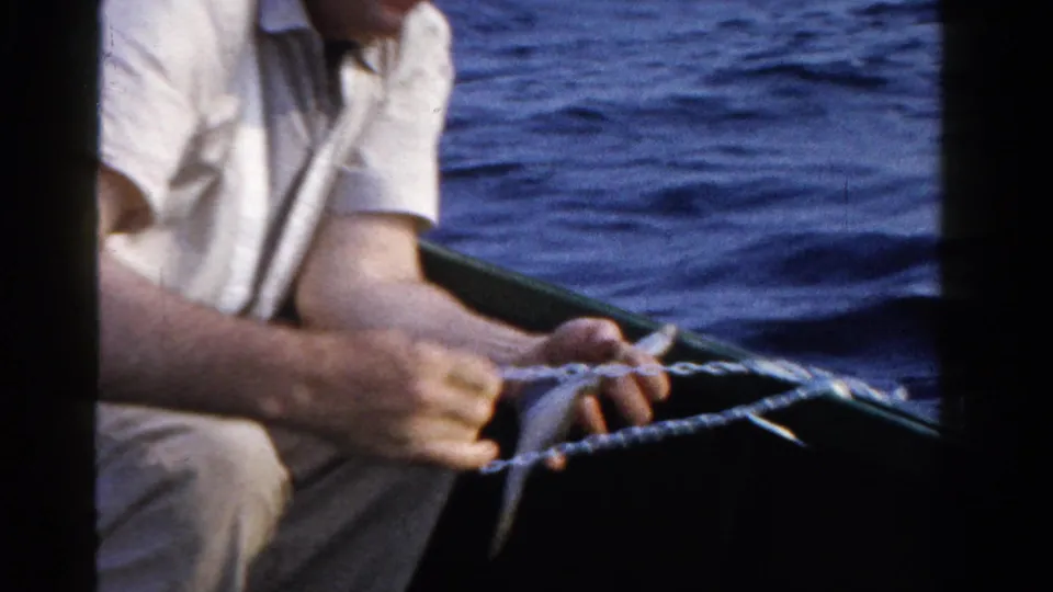 Close 1958 Ontario home-movie frame of hands securing a small fish and line inside a boat with blue water in the background.