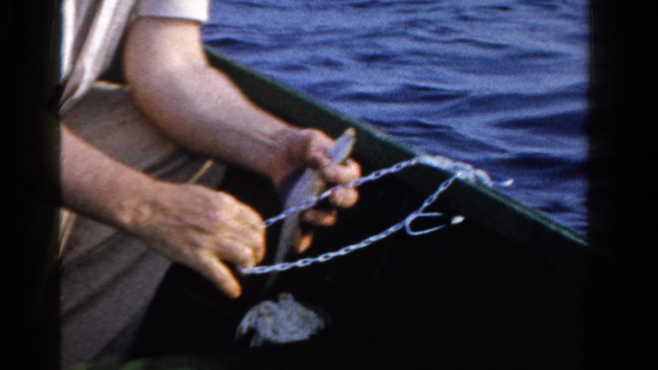 In 1958 Ontario home-movie footage, a man in a light shirt sits in a small boat and handles a fishing line over blue lake water.