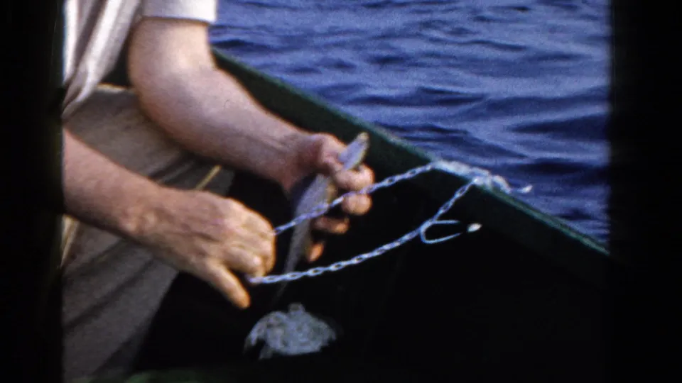 In 1958 Ontario home-movie footage, a man in a light shirt sits in a small boat and handles a fishing line over blue lake water.