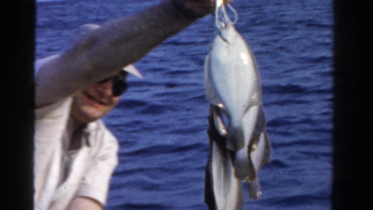 A man in sunglasses aboard a small boat lifts a freshly caught fish above the blue lake water in the supplied 1958 Ontario footage.