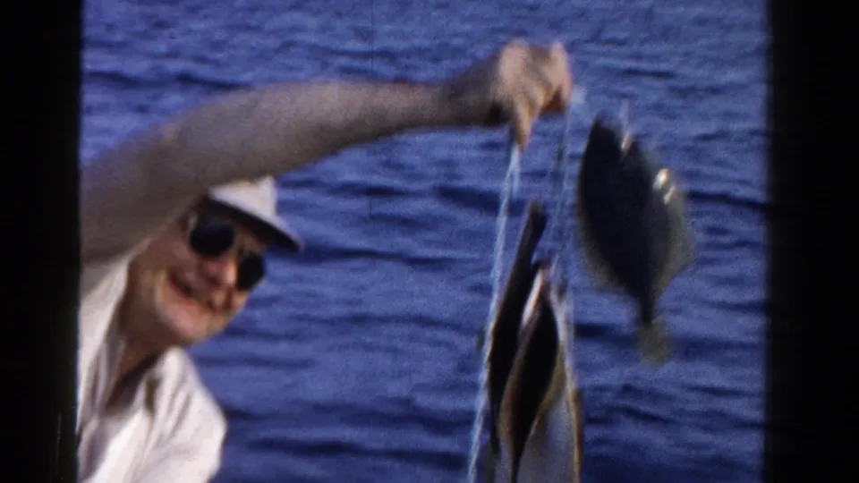The man in sunglasses smiles while lifting a stringer of fish from the boat, with blue lake water filling the background.