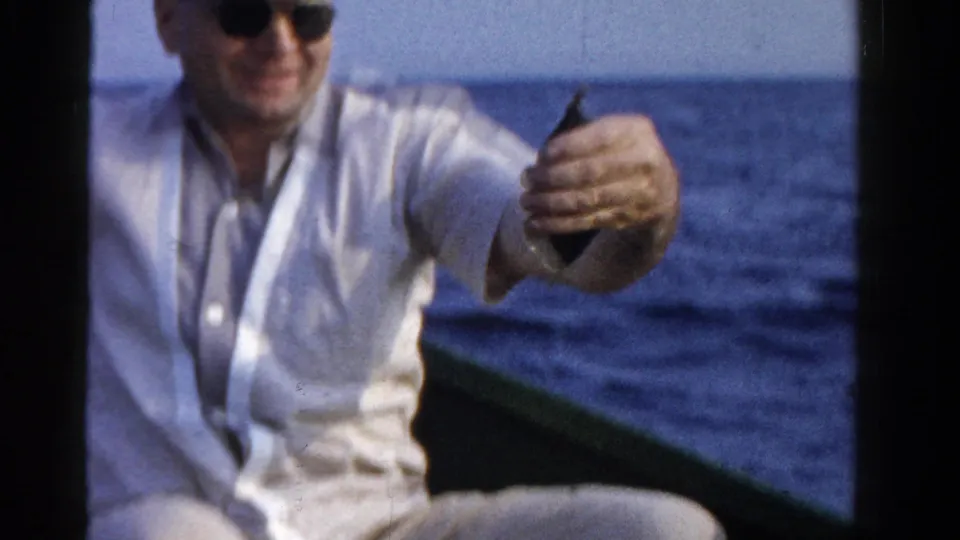 Older man in sunglasses smiles and presents a small fish while seated in a boat on Ontario water in 1958 footage.