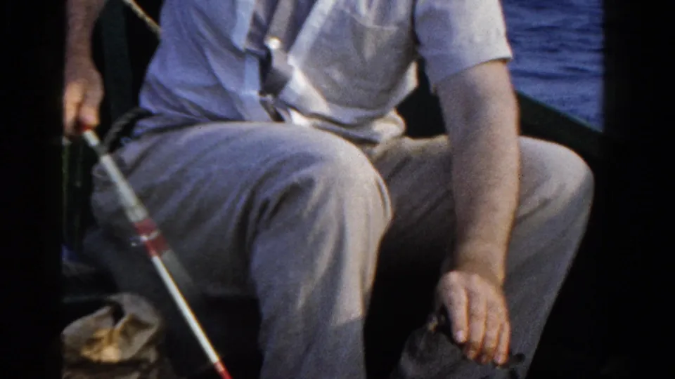 A seated man in light clothing holds a fishing rod in a small boat on Ontario water, with the 1958 home-movie frame cropped at his face.
