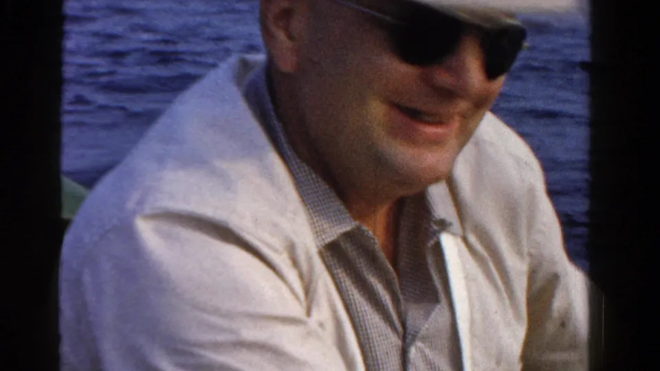 A smiling adult man in sunglasses and a light shirt sits on a boat with open water behind him in 1958 Ontario footage.