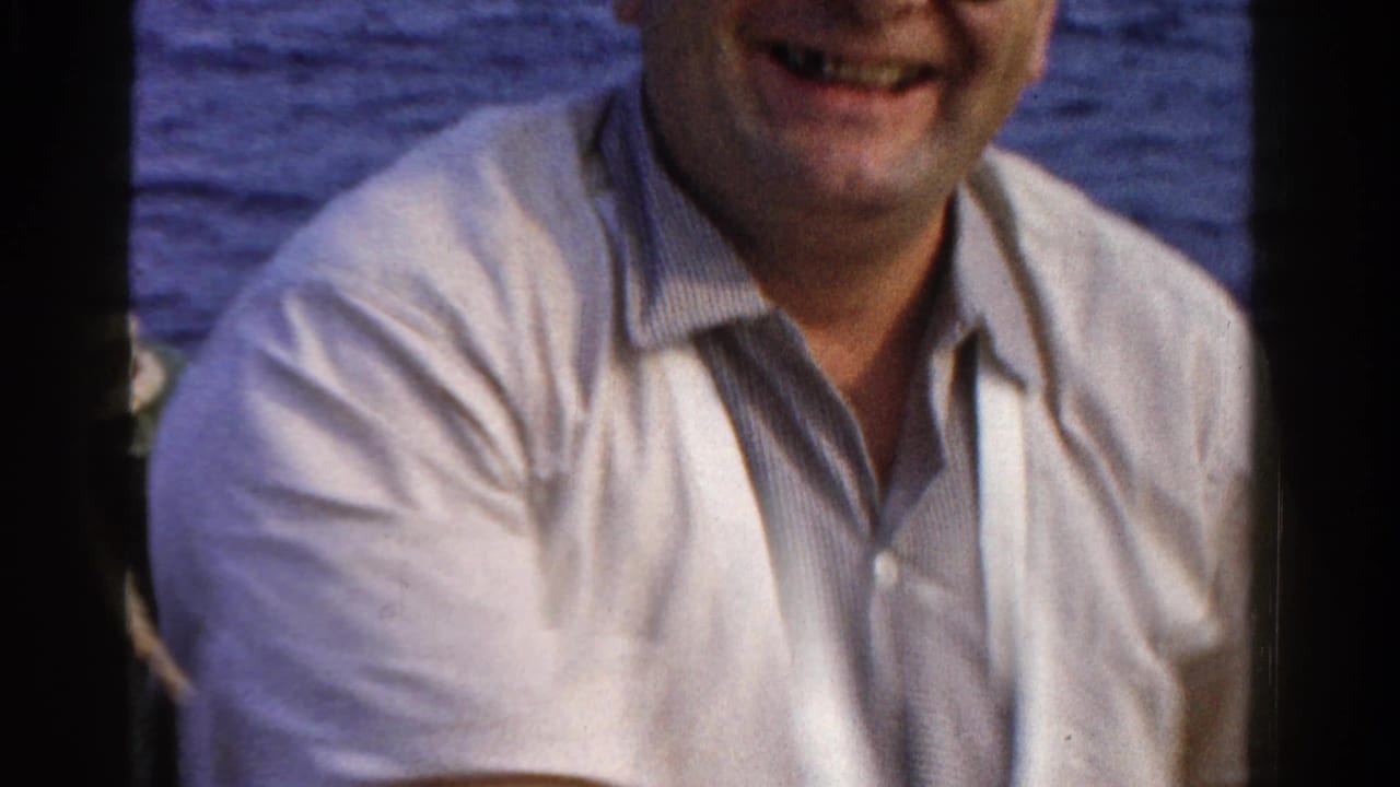 Smiling man in a light shirt seated on a boat with blue water behind him.