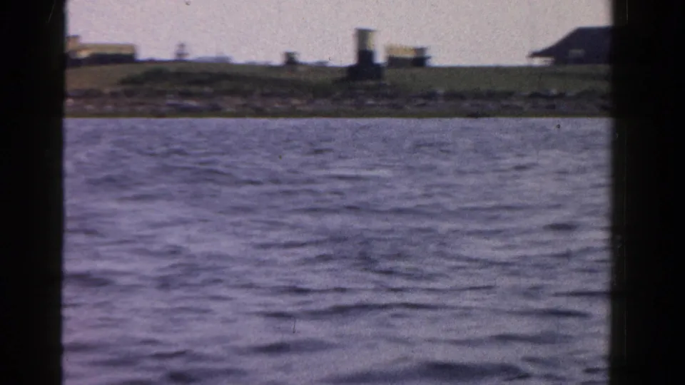 Ontario 1958 shoreline view from the water, with low buildings and greenery visible beyond the waves.