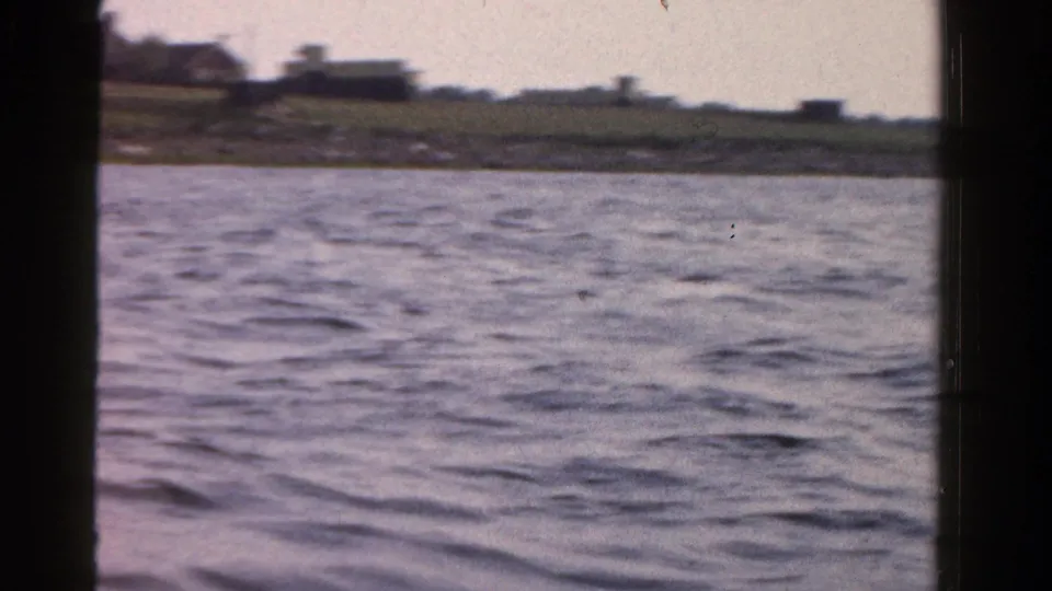 A 1958 Ontario home-movie frame shows water in the foreground and a soft, distant shoreline with low buildings.