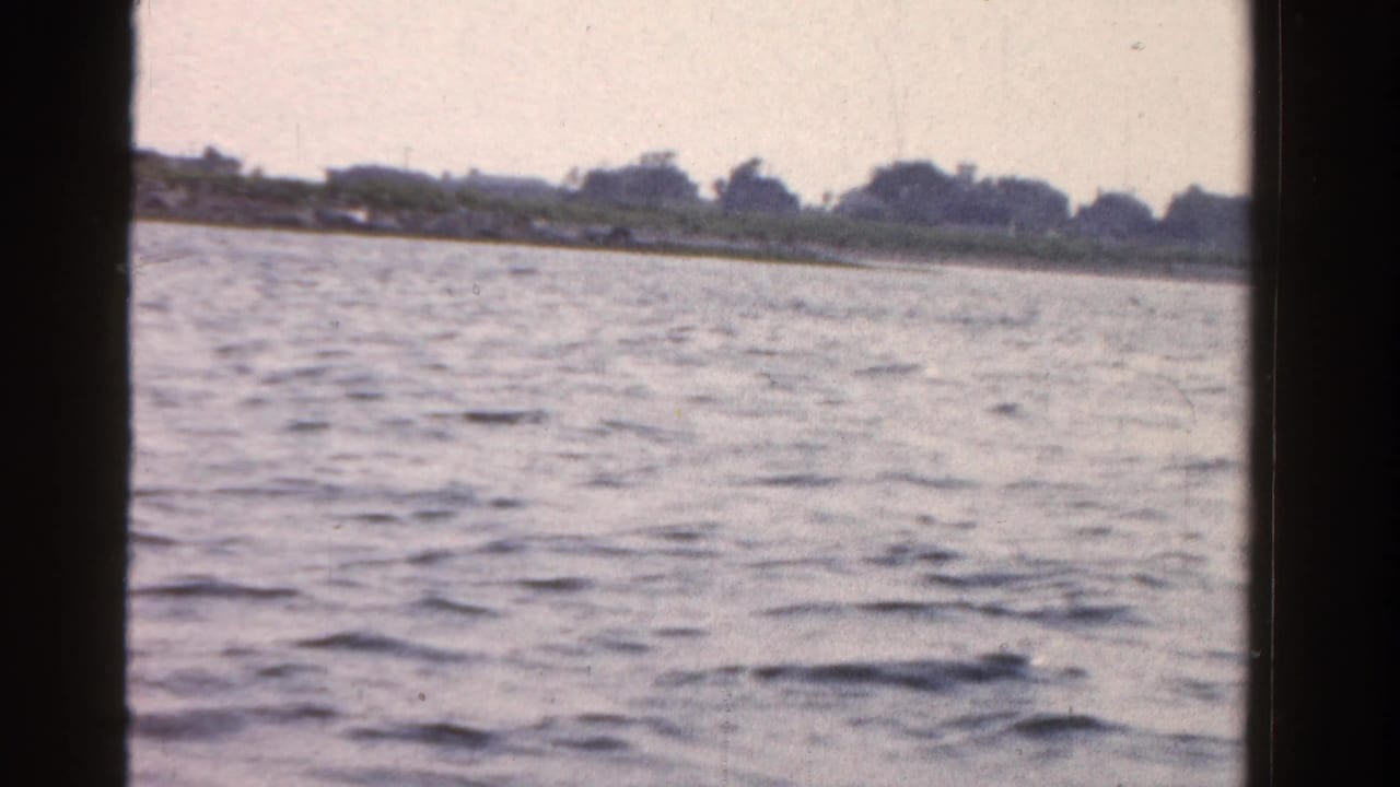 1958 Ontario home-movie frame showing open water, a distant tree-lined shore, and dark window or porthole edges from a boat.