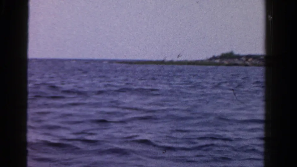 1958 Ontario 8mm frame with blue rippling water, a rocky point, and a small distant structure near the shoreline.