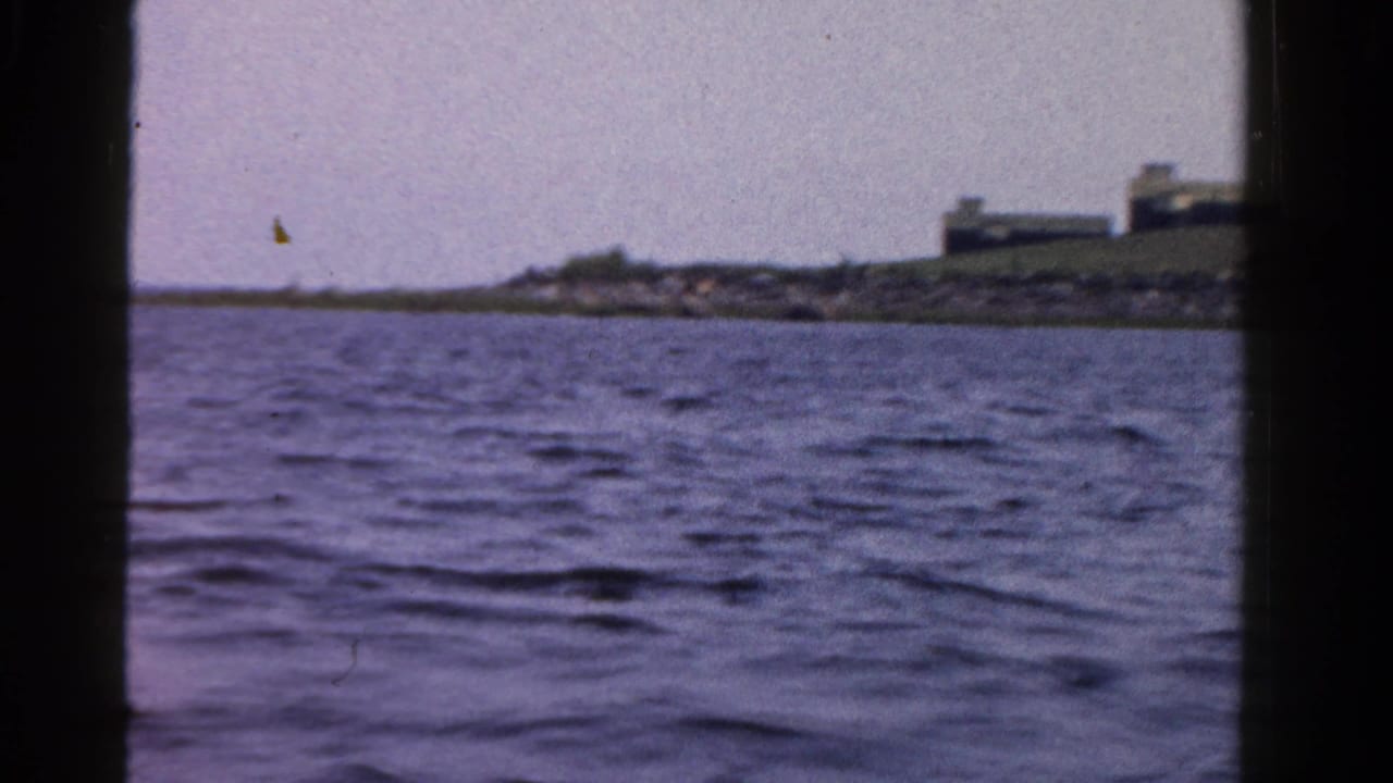 1958 Ontario 8mm frame showing rippled water from a boat, with low buildings and shoreline in the distance.