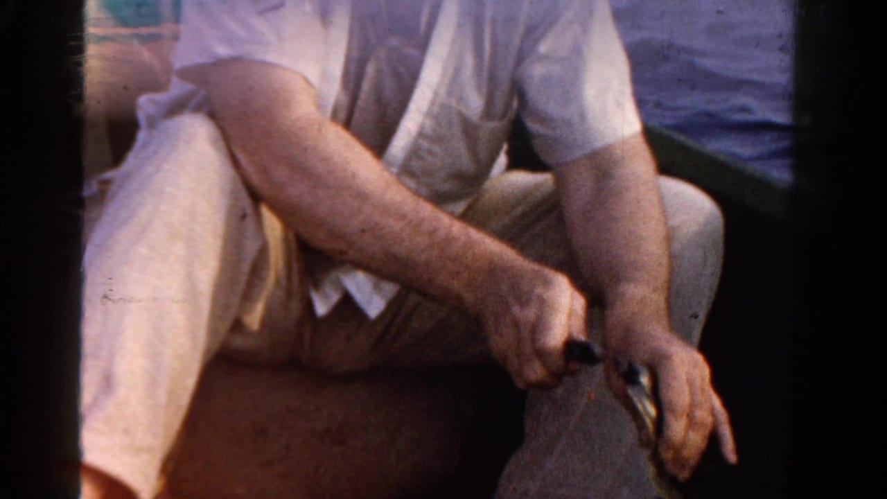 1958 Ontario home-movie frame of a seated adult in light clothing handling fishing gear inside a boat.