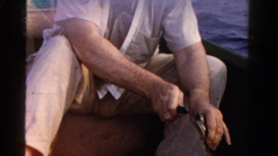 1958 Ontario home-movie frame of a seated adult in light clothing handling fishing gear inside a boat.