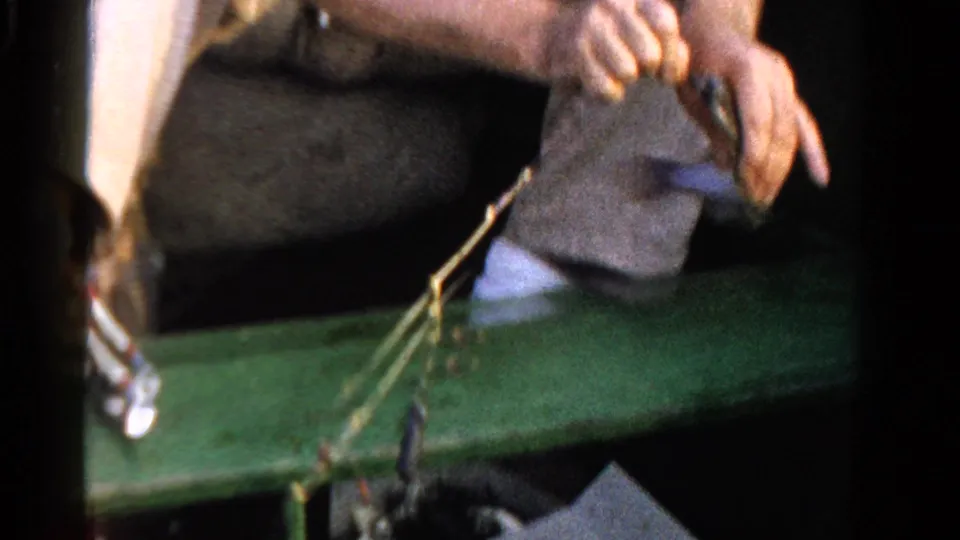 Close home-movie frame of hands managing fishing gear beside a green boat edge, with a small catch or lure visible below.
