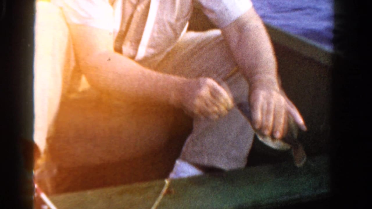 1958 Ontario home-movie frame showing a seated person in light clothing handling fishing gear in a small wooden boat.