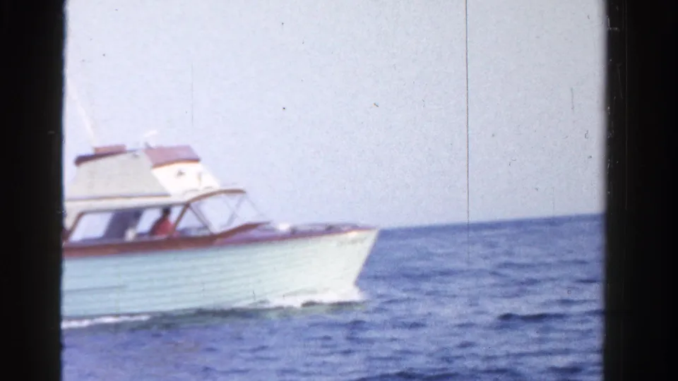 The yacht passes close to the camera frame, with its bow, cabin windows, open water, and pale sky visible.