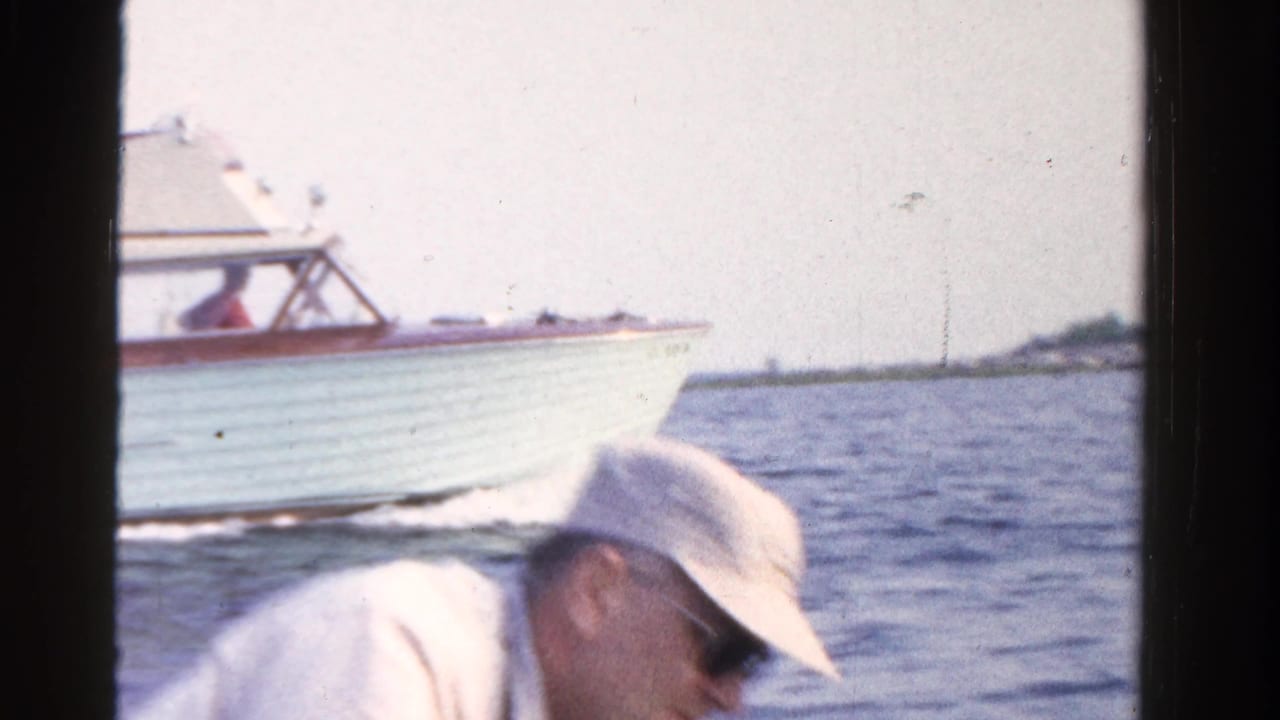 1958 Ontario lake scene with an adult in a white hat and sunglasses in the rowboat foreground as a motorboat passes.