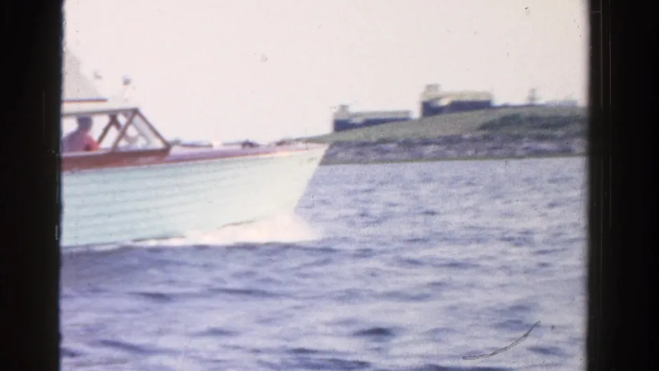 1958 Ontario 8mm frame of a light motorboat crossing lake water with wake and shoreline in the background.