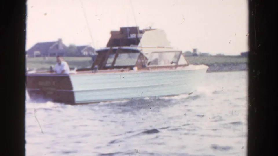 A side view of the vintage motorboat shows its pale hull and wood trim as it passes a low shoreline in Ontario.